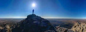 a determined person standing on top of a mountain peak, overlooking a vast, challenging terrain, with a clear blue sky and the sun shining brightly.