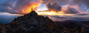 a person standing triumphantly on top of a towering mountain peak, arms raised in victory against a dramatic sunset sky.