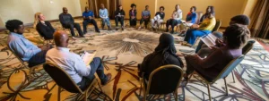 a diverse group of people sitting in a circle, engaged and focused, as they listen attentively to a motivational speaker holding a book titled "you were born rich."